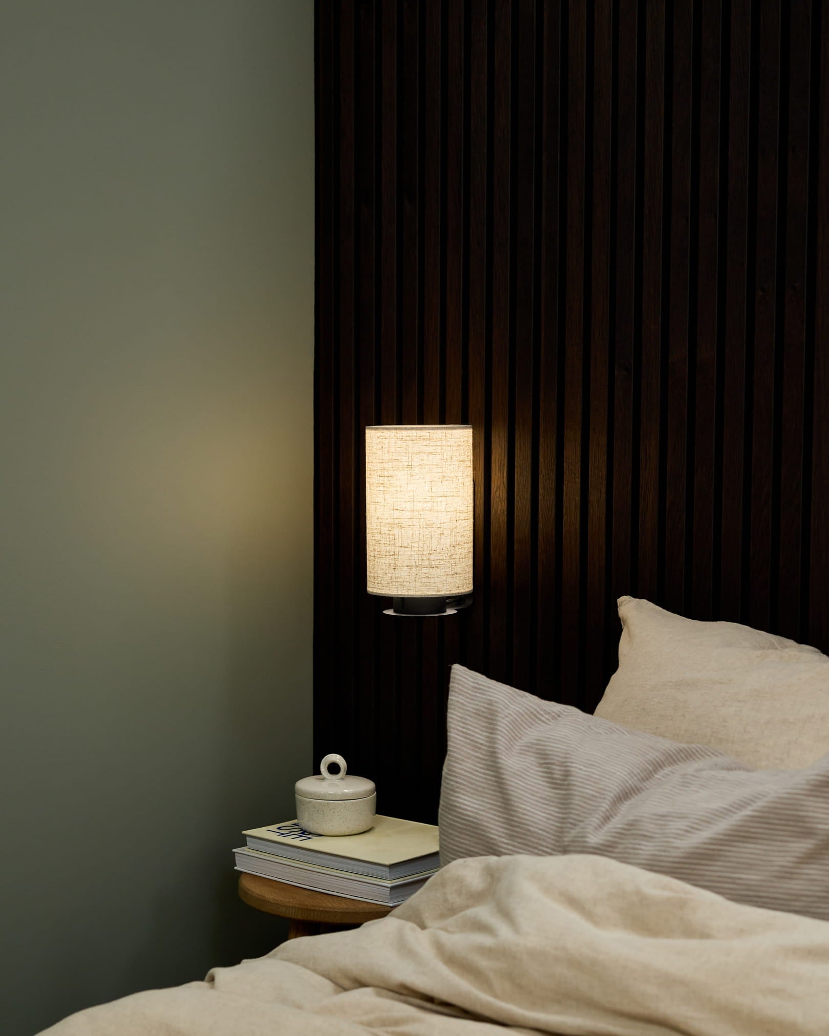 Bedroom corner featuring a WoodUpp Feya Wall Lamp in Black/Beige, beige bedding, and a bedside table with books and a lidded ceramic cup.